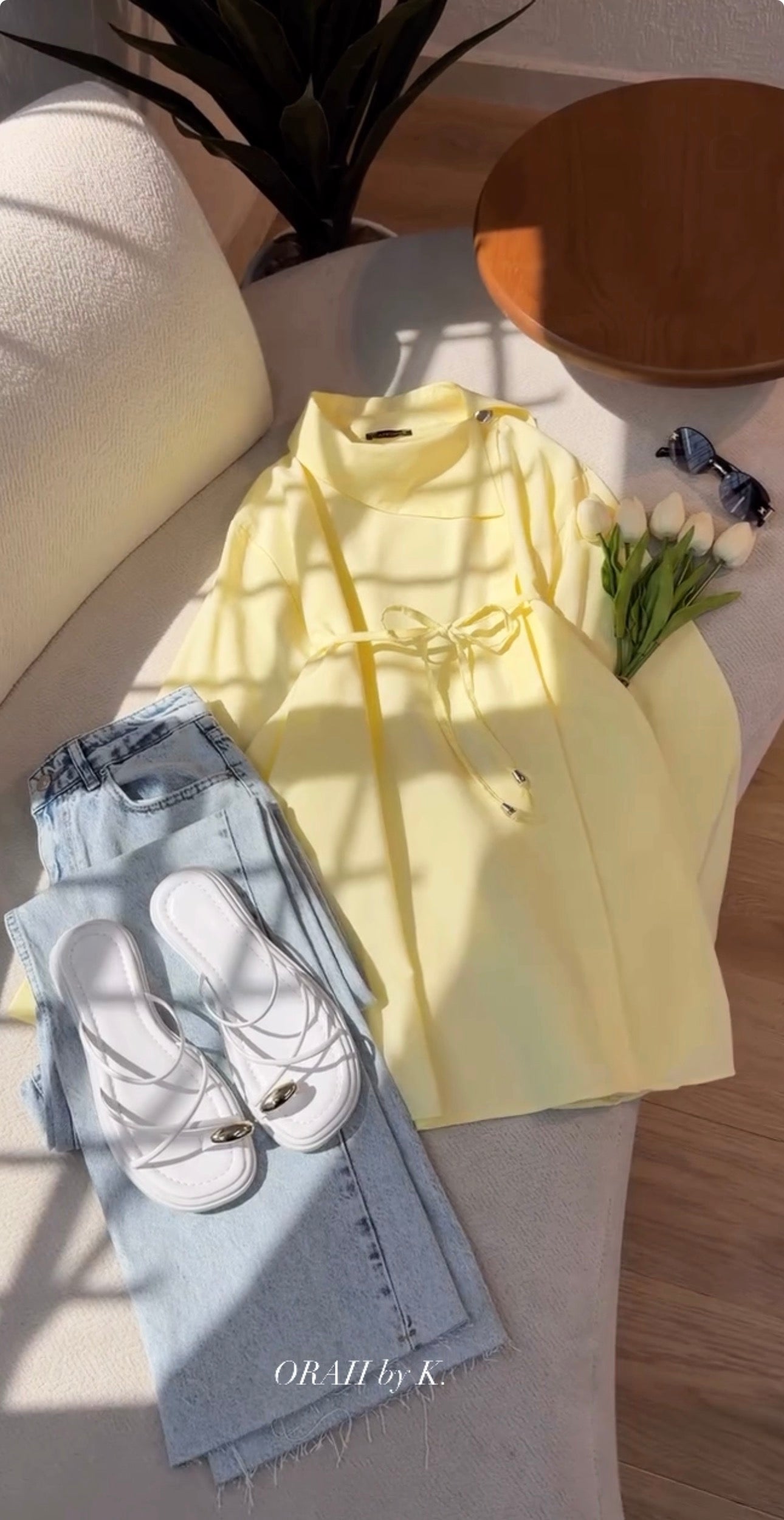 A pale yellow long-sleeve top with a drawstring waist, styled in a flat lay on a light beige sofa alongside light blue jeans, white slide sandals, sunglasses, and a small bouquet of white tulips. The brand name 'ORAH by K.
