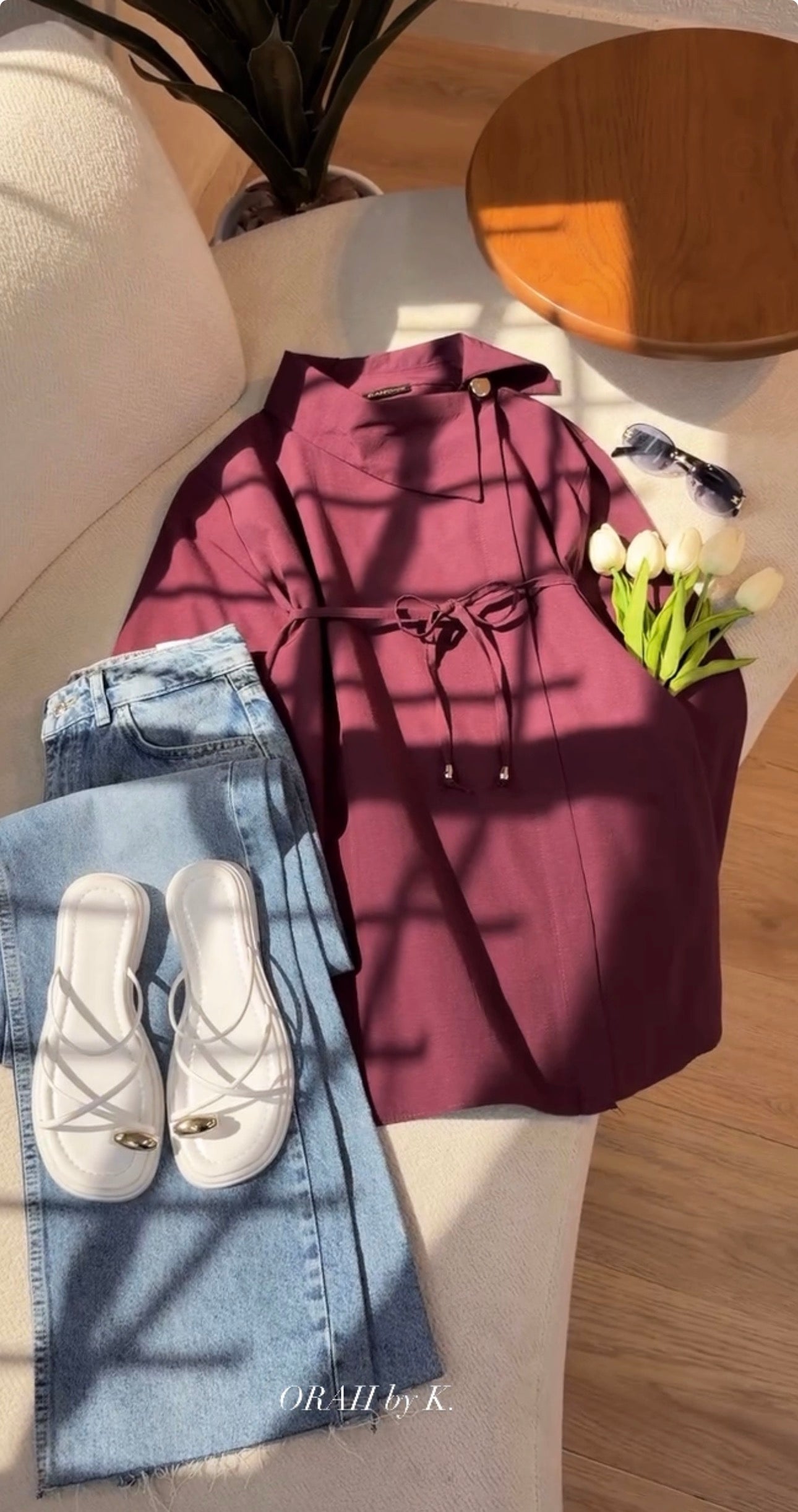 A styled flat-lay of a plum-colored long-sleeve top with a drawstring waist, arranged on a cream sofa alongside light-wash jeans, white slide sandals, white tulips, and a wooden side table in a sunlit room.