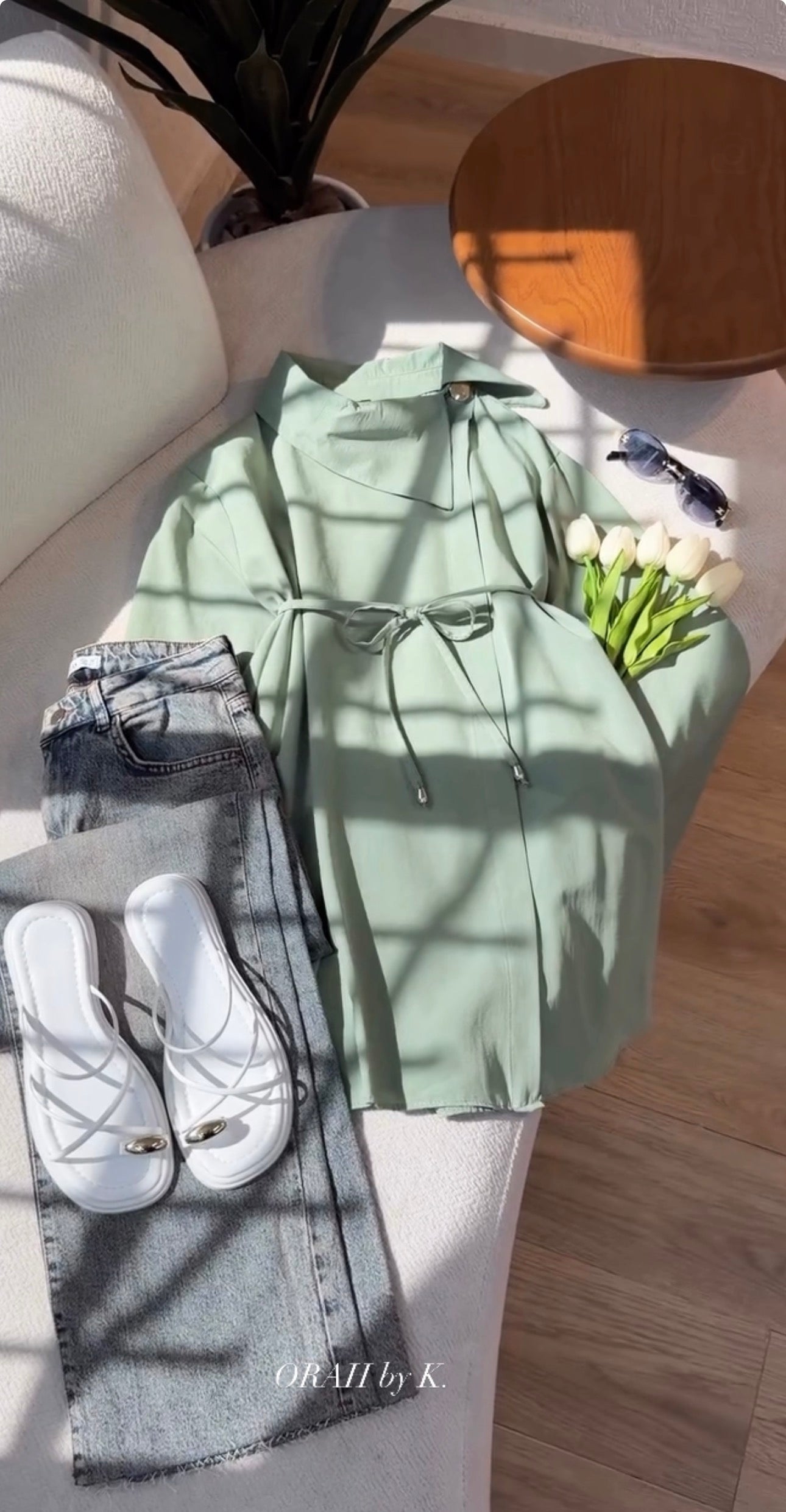 A sage green long-sleeve shirt dress with a drawstring waist, styled in a flat lay with gray jeans, white slip-on shoes, sunglasses, and white tulips on a white sofa with dappled sunlight.